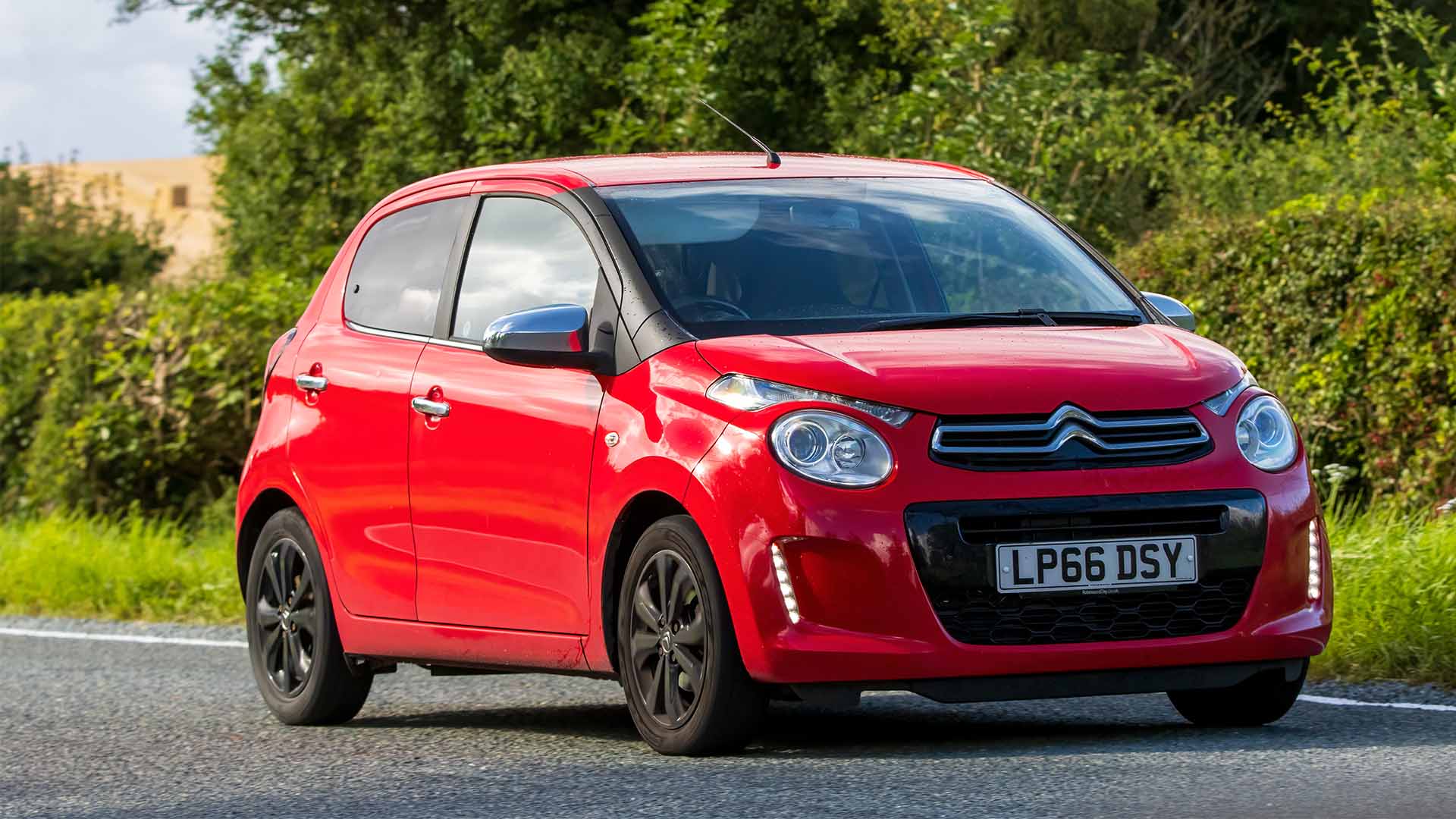 Red Citroën C1 driving past trees and bushes